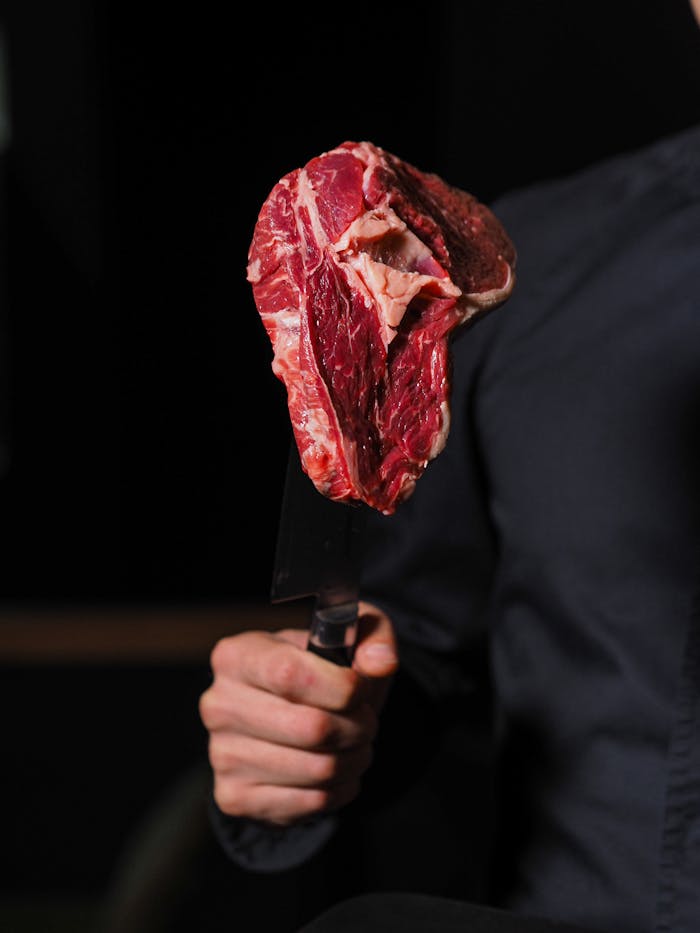A person holds a raw steak on a knife, ready for cooking. Dark indoor setting.